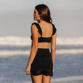 Back view of Black Bella Crop Top showcasing ruffled shoulder straps on a model against a beach background
