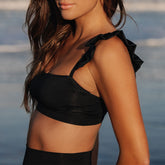 Close-up side view of a model wearing the black Bella Crop Top with ruffled shoulder straps against a blue outdoor background