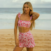 Front view of the pink and white floral Athena Bella Crop Top showcasing a square neckline and ruffled straps on a beach