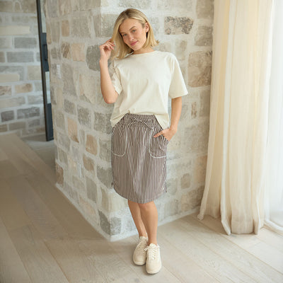 Woman wearing a white top and striped skirt standing in a room with stone walls and a curtain.