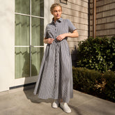 Woman wearing a long, navy blue gingham dress standing outside a near a window and bushes.