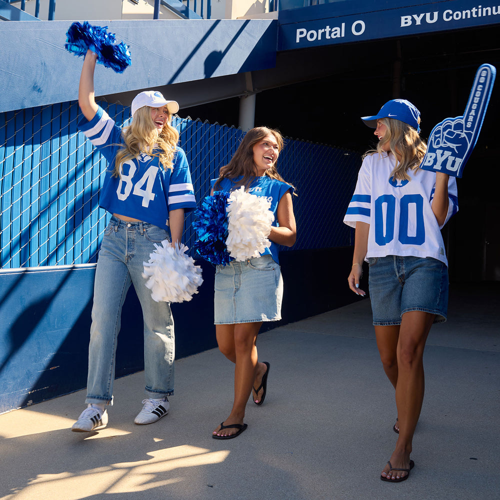 Three women in BYU apparel with pom-poms outside a stadium entrance, including a royal jersey with number 84