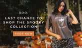 A woman wearing a dark brown graphic t-shirt with a skull design and 'Mostly Tricks, Some Treats' text, paired with black and white patterned shorts, standing in front of a pumpkin display with promotional text "Last chance to shop the spooky collection"